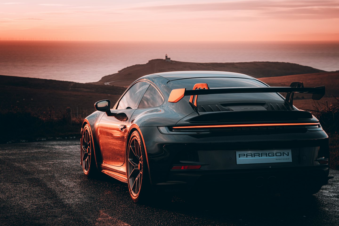 Dark grey sports car with orange spoiler parked on coastal road.